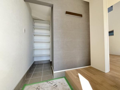 玄関は落ち着いた色合いのタイル仕様で清潔感があります。奥には可動式の棚が設けられ使い勝手が良いです。隣接する壁面にはアクセントとなる照明が設置されています。