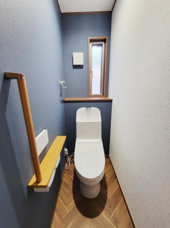 ブルーの壁面と木目調の床が調和したトイレです。小窓から自然光が差し込み空間を明るくしています。手すりが設置されており安心です。収納棚が備え付けられているため整理しやすいです。