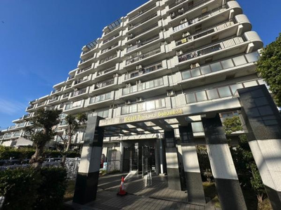 堂々とした佇まいが印象的なマンションです。エントランスにはしっかりとした門構えがあります。日差しを受けて明るい外観が映えています。周辺には緑も見られます。