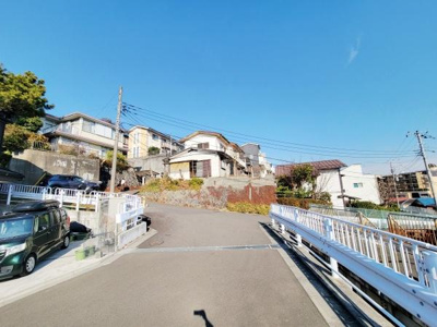 開放的な青空が広がる住宅街の風景です。坂道沿いの立地で見晴らしが良いイメージです。周囲には整った街並みが広がっています。道路が広めで車の出し入れもスムーズにできます。