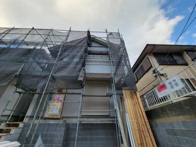 建物全体が足場とネットでしっかりと覆われており工事中の様子が確認できます。外壁や窓が見え建物の構造がはっきりと伝わります。現地の雰囲気や敷地の広さも感じられる写真です。