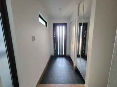 玄関はシンプルなデザインで明るい光が差し込みます。大きな鏡付きの収納がありスッキリとした印象です。ガラス入りのドアがモダンな雰囲気を演出しています。落ち着いた色合いの床タイルが特徴的です。