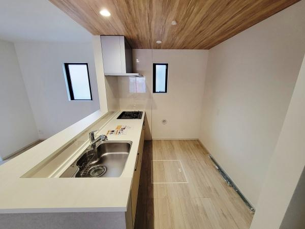 天井の木目と床の色合いが調和したキッチンです。二つの窓から自然光が入り明るい空間となっています。ワイドなカウンターとシンクが使いやすい設計です。コンロやシンクがまとめられており動線がシンプルです。