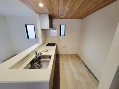 天井の木目と床の色合いが調和したキッチンです。二つの窓から自然光が入り明るい空間となっています。ワイドなカウンターとシンクが使いやすい設計です。コンロやシンクがまとめられており動線がシンプルです。