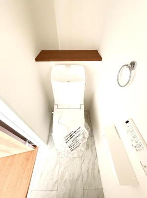 白を基調とした清潔感のあるトイレです。床は大理石調で空間を明るく演出します。収納に便利な棚も備え付けられています。シンプルなデザインが特徴です。