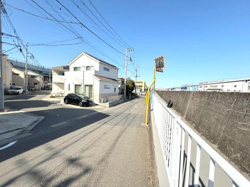 前面道路は広めで見通しが良いです。周囲には住宅が立ち並び明るい印象の現地です。電柱やフェンスもしっかり整備されています。道路沿いには歩道もあり安全性が感じられます。晴れた日の空が広がり開放的です。