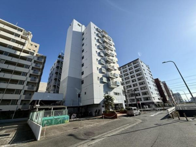 白を基調とした外観が印象的なマンションです。周囲にもマンションが建ち並ぶエリアで街並みに調和しています。道路沿いでアクセスがしやすい構造です。建物の敷地には植栽も施されています。