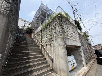 道路から階段を上がった先に敷地が広がる高台の建物です。堅牢な石垣とコンクリートの外観が印象的です。車庫スペースも確保されており利便性があります。周囲に緑の植栽があしらわれています。