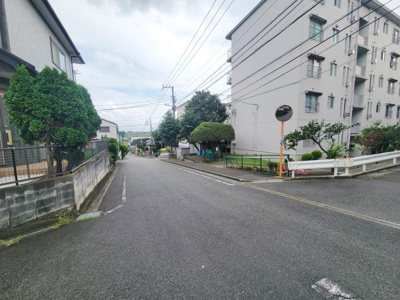 住宅街の一角に位置し整備された道路が広がっています。周囲には緑が多く落ち着いた雰囲気が感じられます。白いマンションや戸建てが並び開放感のある通りです。道路幅が広く車や自転車も通りやすい環境です。
