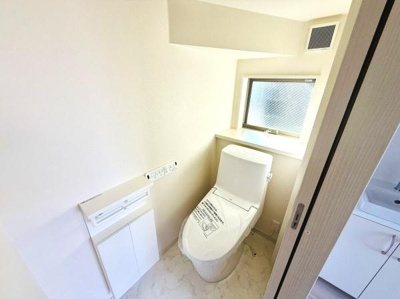 窓があり換気がしやすいトイレ空間です。シンプルなデザインで清潔感があります。壁付けのペーパーホルダーや収納も設置されています。入り口は引き戸仕様で使いやすい設計になっています。