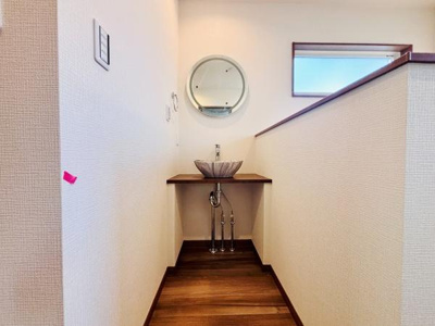 シンプルなデザインの洗面スペースです。丸型のミラーとコンパクトな洗面台が特徴です。木目調の床が空間に温かみを添えています。上部には窓もあり明るさを感じます。