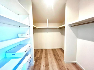 棚やハンガーパイプが備え付けられており整理しやすい収納スペースです。明るい照明で室内が見渡しやすくなっています。木目調の床が落ち着いた雰囲気を演出しています。