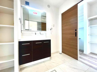 洗面台と収納棚がすっきりとレイアウトされています。明るく清潔感のある空間が広がっています。大きな鏡で身支度も快適に行えます。木目調の扉が落ち着いた印象を与えています。ゆとりある洗面所スペースです。