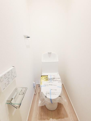 シンプルで清潔感のあるデザインのトイレです。壁面に設置された操作パネルが便利です。床材の色合いが明るい空間を演出します。ペーパーや小物類を置けるガラス棚も備え付けられています。