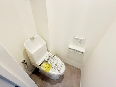 シンプルな内装のトイレです。清潔感のある空間が特徴です。壁掛けタイプのペーパーホルダーが備え付けられています。操作パネルも視認性の高い位置に設置されています。床材と壁面の色合いが調和しています。