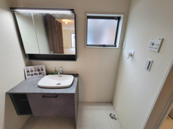 シンプルでスタイリッシュな洗面台が設置されています。大きな鏡と収納スペースを備えており日常使いに便利です。窓からの自然光が明るさをプラスしています。機能的なレイアウトが魅力の洗面所です。