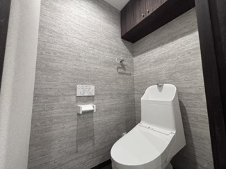 グレーの壁面とダークブラウンの収納棚が洗練された印象を与えるトイレです。温水洗浄機能付きで快適にご利用いただけます。手洗い一体型のタンクで使い勝手も良好です。