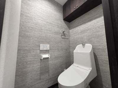 グレーの壁面とダークブラウンの収納棚が洗練された印象を与えるトイレです。温水洗浄機能付きで快適にご利用いただけます。手洗い一体型のタンクで使い勝手も良好です。