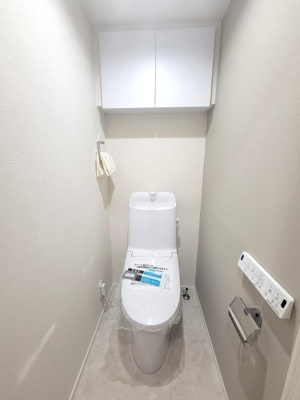 清潔感のあるシンプルなトイレです。上部に収納棚が設置されており整理整頓がしやすい空間です。壁付けのペーパーホルダーや操作パネルも設置されています。全体的に明るい印象の内装です。