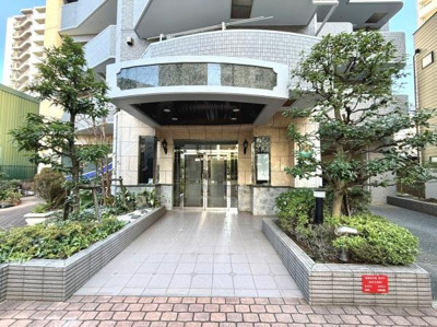 植栽が施されタイル張りのアプローチが印象的なエントランスです。ガラス扉で明るい雰囲気が漂います。重厚感のある庇やシンプルながらも洗練された外観が目を引きます。清潔感を感じるデザインが特徴です。